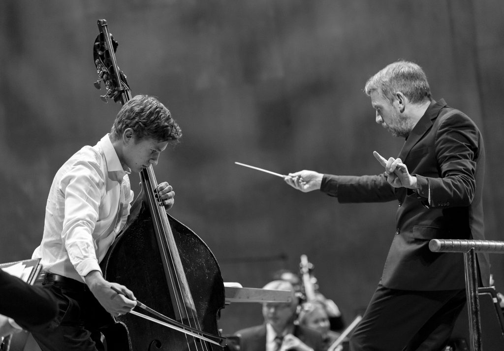 Ung kille spelar cello, framför dirigerar en man