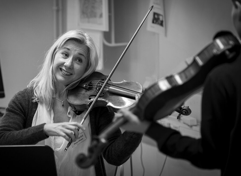 En glad kvinna spelar fiol och ler samtidigt. Framför henne står en person som också spelar fiol.